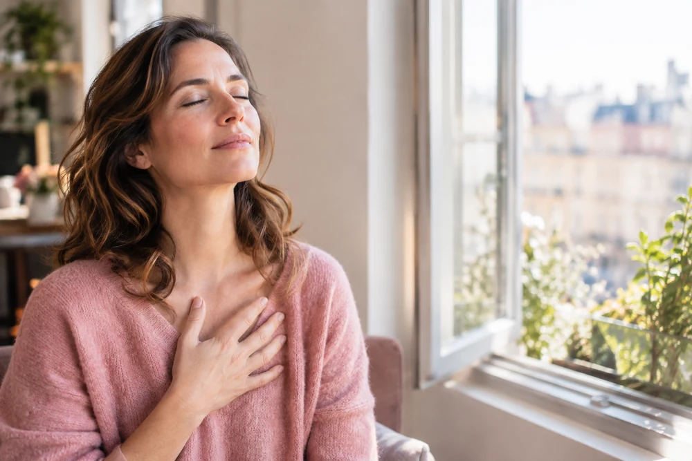 Calmer l'attachement anxieux : une femme respire les yeux fermés, main sur la poitrine, près d'une fenêtre ouverte