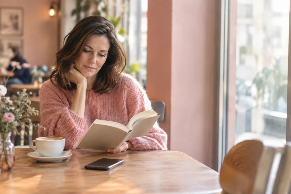 Il regrette la rupture : une femme sereine lit un livre dans un café, son téléphone posé face contre la table